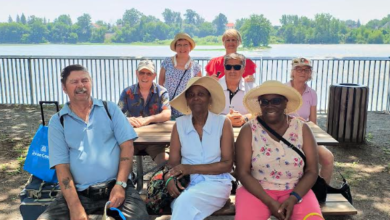 Group of seniors posing at a lakeside park picnic table under leafy trees, smiling with a sunny day by the water.