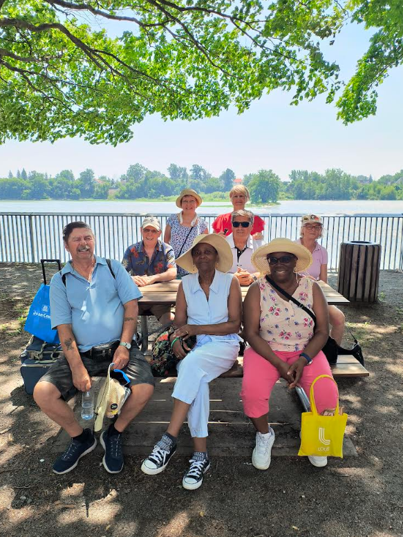 Club de marche les deux berges Média Laval Group of seniors posing at a lakeside park picnic table under leafy trees, smiling with a sunny day by the water.