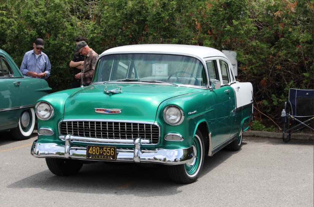 Vintage green and white Chevrolet at a car show with people examining it in the background, chrome bumper and grille in focus.