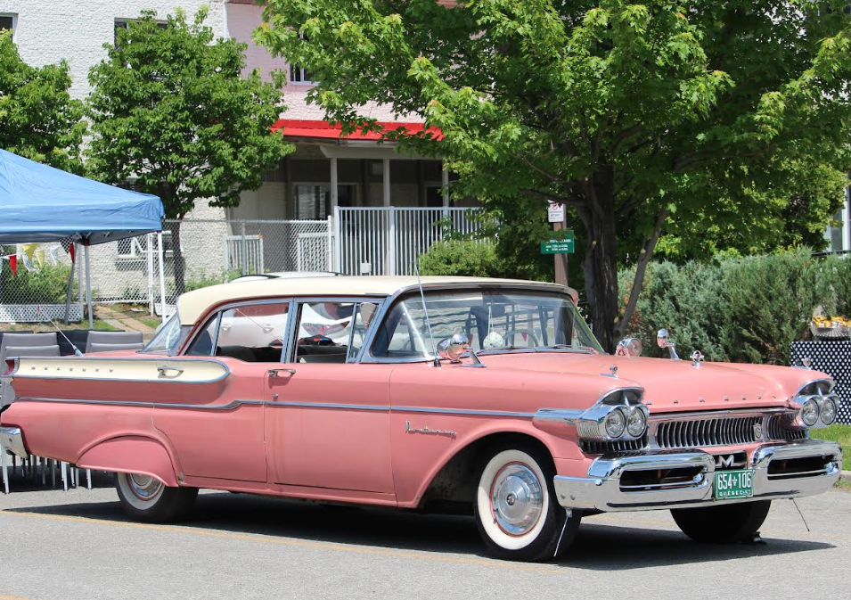 Classic pink 1950s car with chrome trim and whitewall tires parked on a sunny street at a car show under a blue canopy.