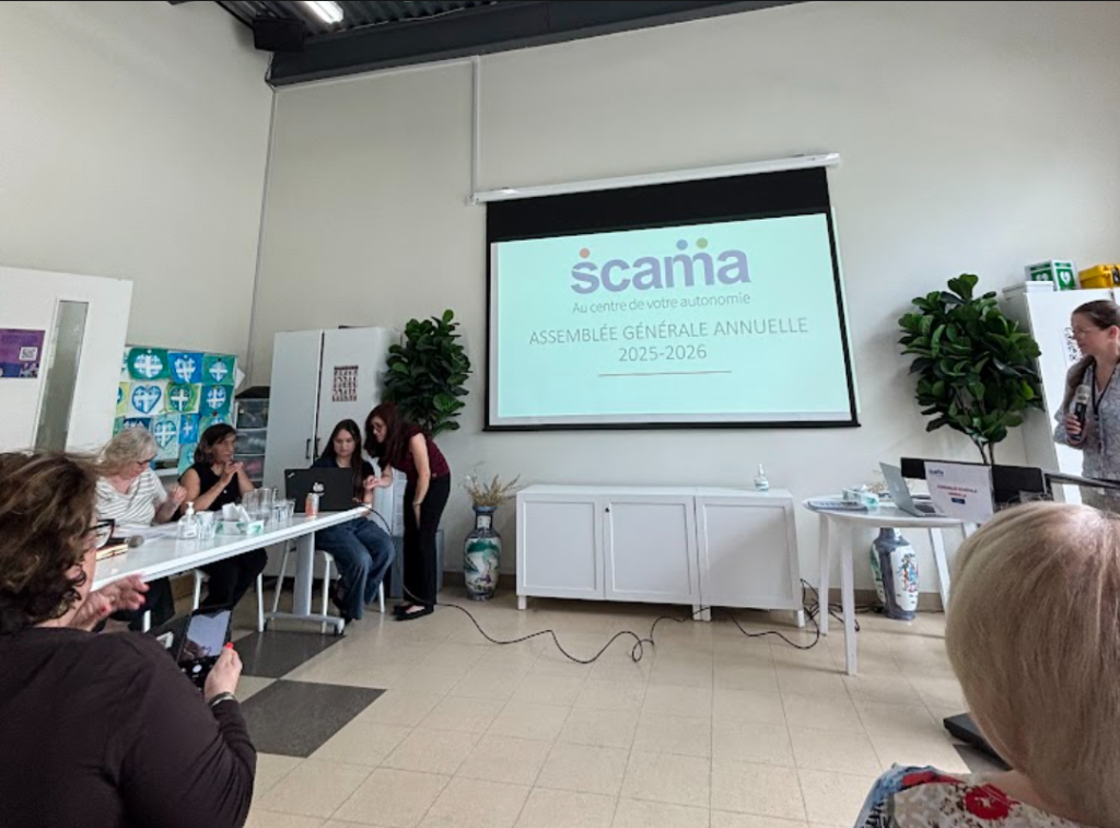 AGA Centre Scama 2026 Média Laval Group of people seated around a long table in a bright meeting room, watching a projector screen displaying a slide with a'scania' logo and French text.