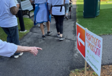 Group of people in white shirts walk along a paved path at an outdoor event, holding a flag with a sign that reads 'Marchons pour la Maison'.