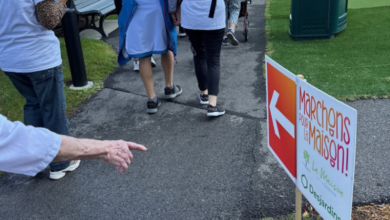 Group of people in white shirts walk along a paved path at an outdoor event, holding a flag with a sign that reads 'Marchons pour la Maison'.
