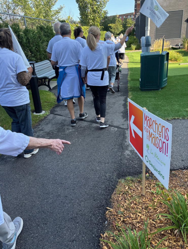 Cinquième édition Maison sérénité Média Laval Group of people in white shirts walk along a paved path at an outdoor event, holding a flag with a sign that reads 'Marchons pour la Maison'.