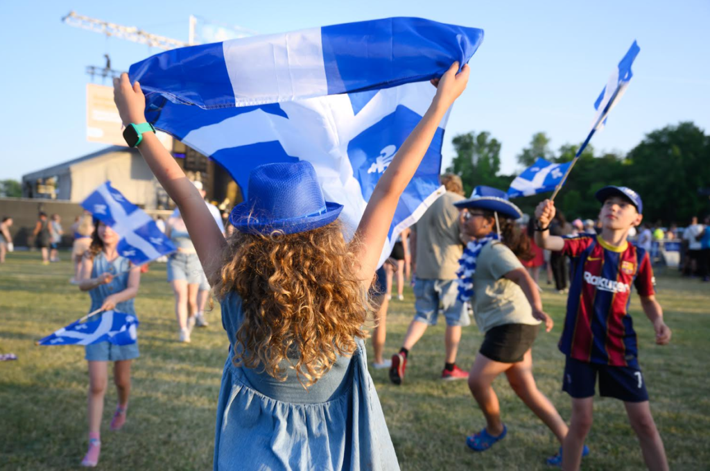 Children at an outdoor event waving blue and white flags, wearing blue hats and casual outfits.