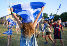 Children at an outdoor event waving blue and white flags, wearing blue hats and casual outfits.