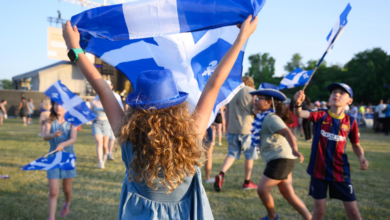 Children at an outdoor event waving blue and white flags, wearing blue hats and casual outfits.