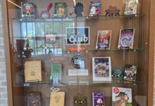 Glass-front book display case with shelves of colorful children's books and props, including magnifying glass and hat silhouettes, centered around a 'Club' sign.