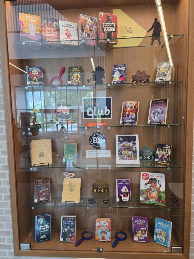 Biblio Club Laval Média Laval Glass-front book display case with shelves of colorful children's books and props, including magnifying glass and hat silhouettes, centered around a 'Club' sign.