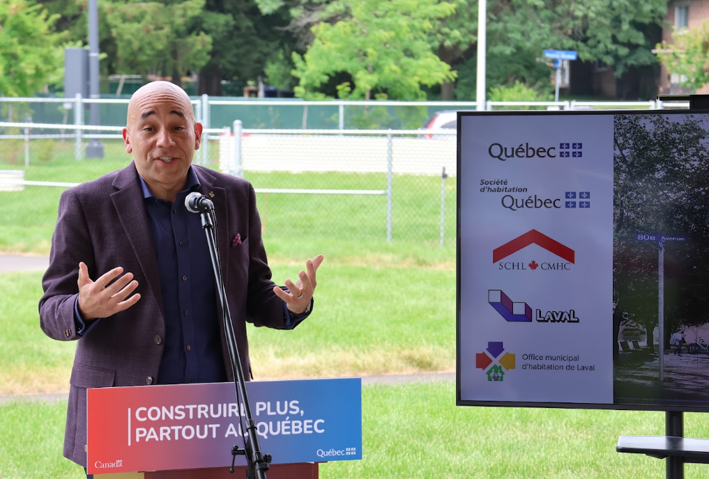 Man in a purple blazer speaks at an outdoor podium with a 'Construire plus, partout au Québec' banner and a screen showing Quebec logos nearby.