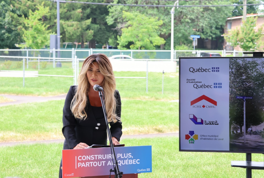 Woman in a black blazer speaks into a microphone at an outdoor press event, standing behind a podium with a Quebec-themed banner.