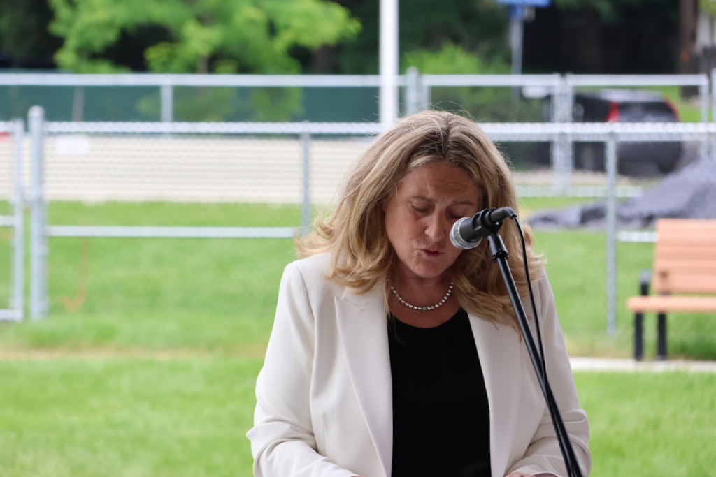 Woman in a white blazer speaks into a microphone outdoors at a park event, with green grass and a fence behind her.