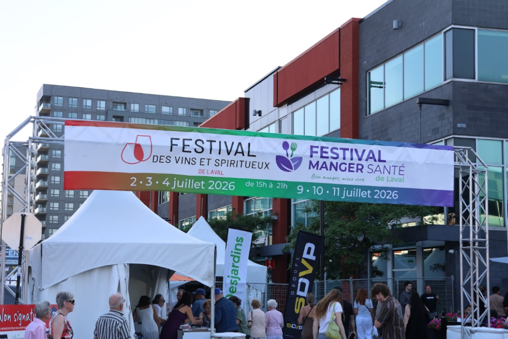 Festival vins et spiritueux de Laval 2026 Média Laval Outdoor festival entrance with a large colorful banner advertising wine, spirits and health-food events in Laval; crowd and white tents beneath.
