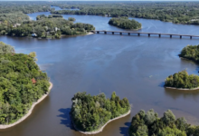 Aerial view of a wide river with tree-covered islands and a long bridge crossing the water. At center, a large island cluster creates a curved inlet.