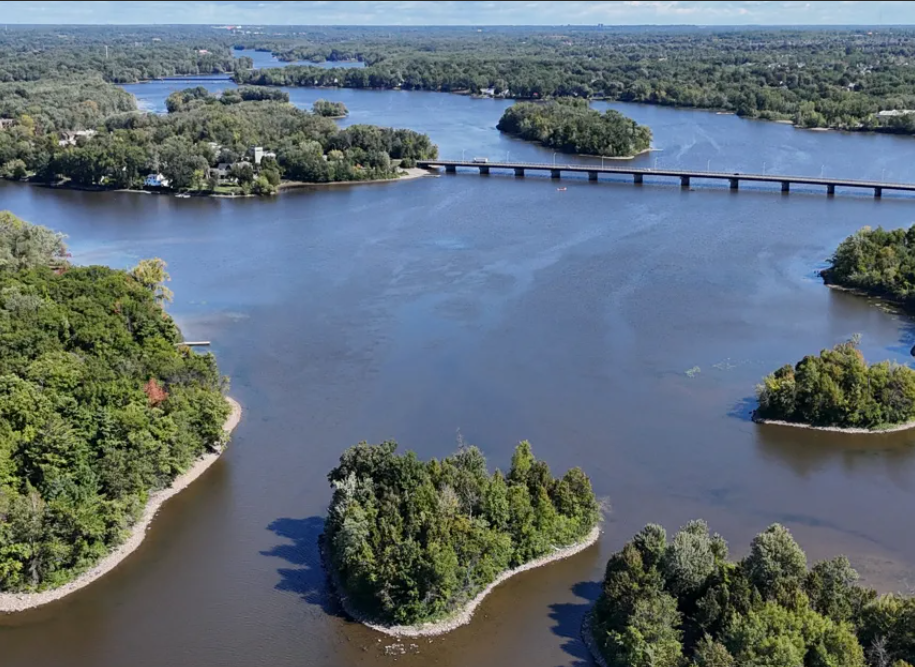 Parc de la RivièredesMilleÎles Média Laval Aerial view of a wide river with tree-covered islands and a long bridge crossing the water. At center, a large island cluster creates a curved inlet.