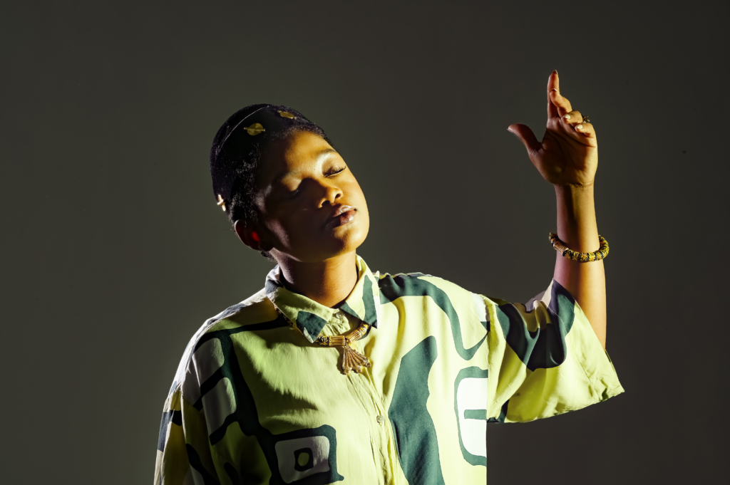 Person with eyes closed raising their right arm in a dance-like pose against a dark background, wearing a yellow-green camouflage shirt and bracelets.