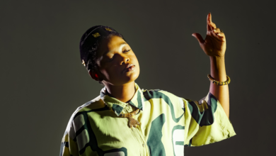 Person with eyes closed raising their right arm in a dance-like pose against a dark background, wearing a yellow-green camouflage shirt and bracelets.
