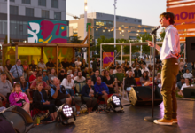 Male comedian on a stage with microphone in front of a large outdoor audience at dusk, colorful murals in the background.
