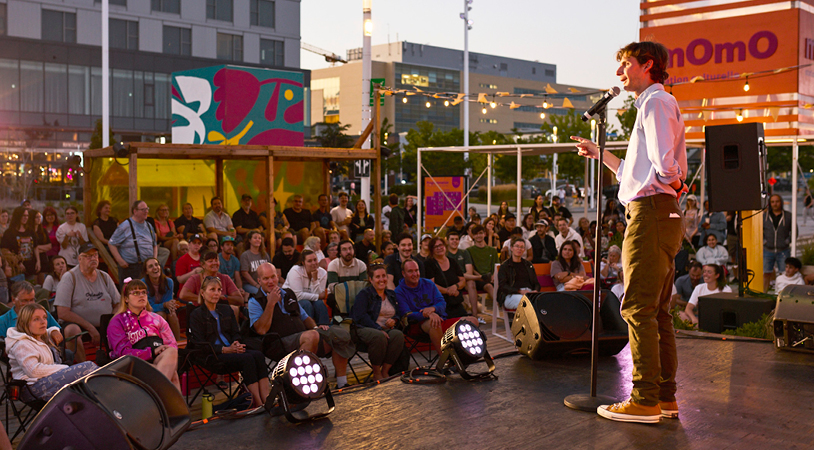 Male comedian on a stage with microphone in front of a large outdoor audience at dusk, colorful murals in the background.