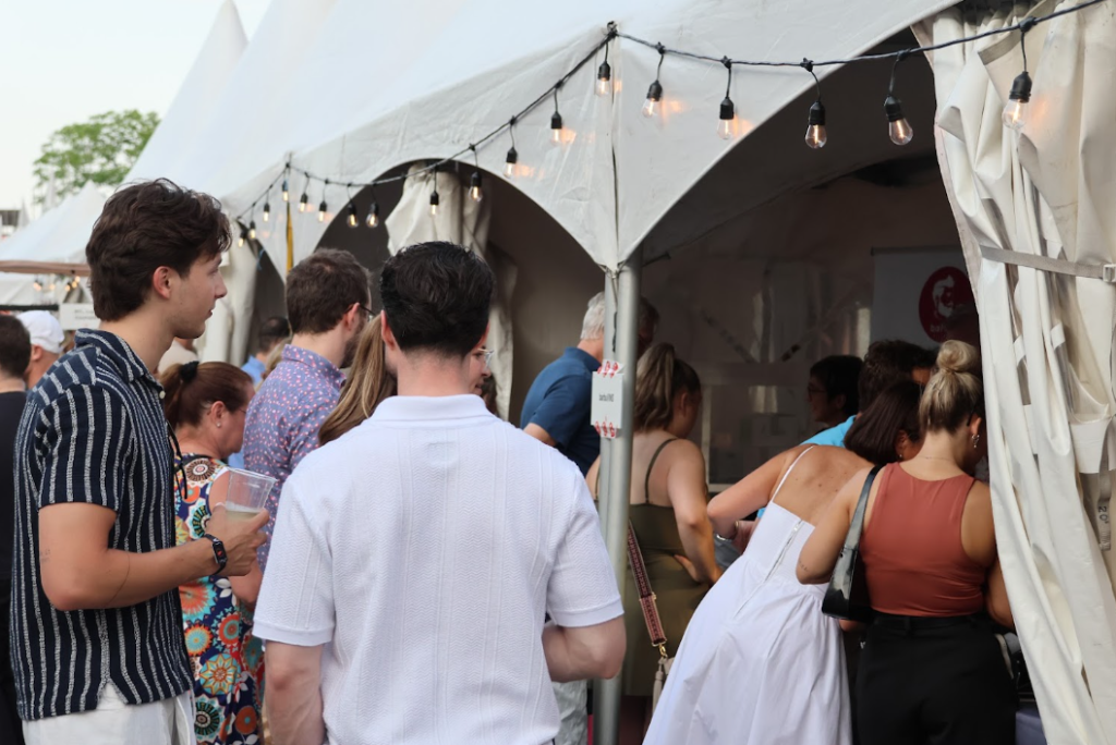 image Média Laval People line up at vendor tents at an outdoor festival, with string lights overhead.