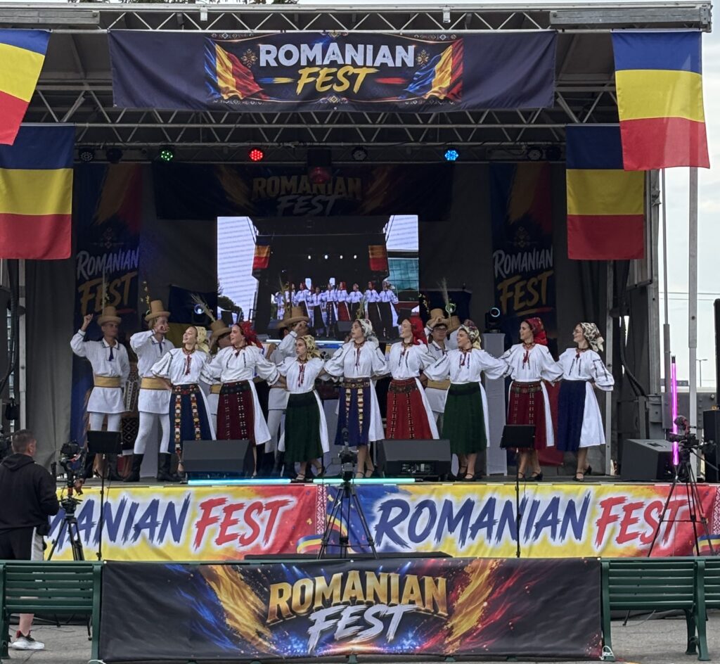 Stage performance at Romanian Fest with dancers in traditional folk costumes and Romanian flags on both sides of the banner.