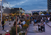 Outdoor evening concert in a plaza with a large crowd, stage at far end, surrounding shops and tents.