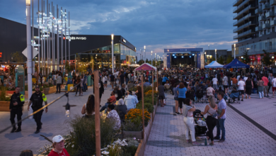 Outdoor evening concert in a plaza with a large crowd, stage at far end, surrounding shops and tents.