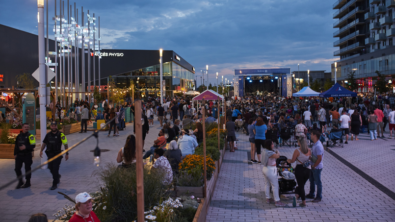 Outdoor evening concert in a plaza with a large crowd, stage at far end, surrounding shops and tents.