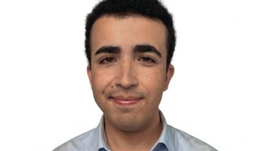 Portrait of a young man with short black hair in a light blue shirt, facing the camera against a white background.