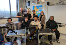 Group of eight people in a classroom recording studio with microphones and audio equipment, posing for a photo.