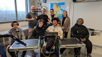 Group of eight people in a classroom recording studio with microphones and audio equipment, posing for a photo.