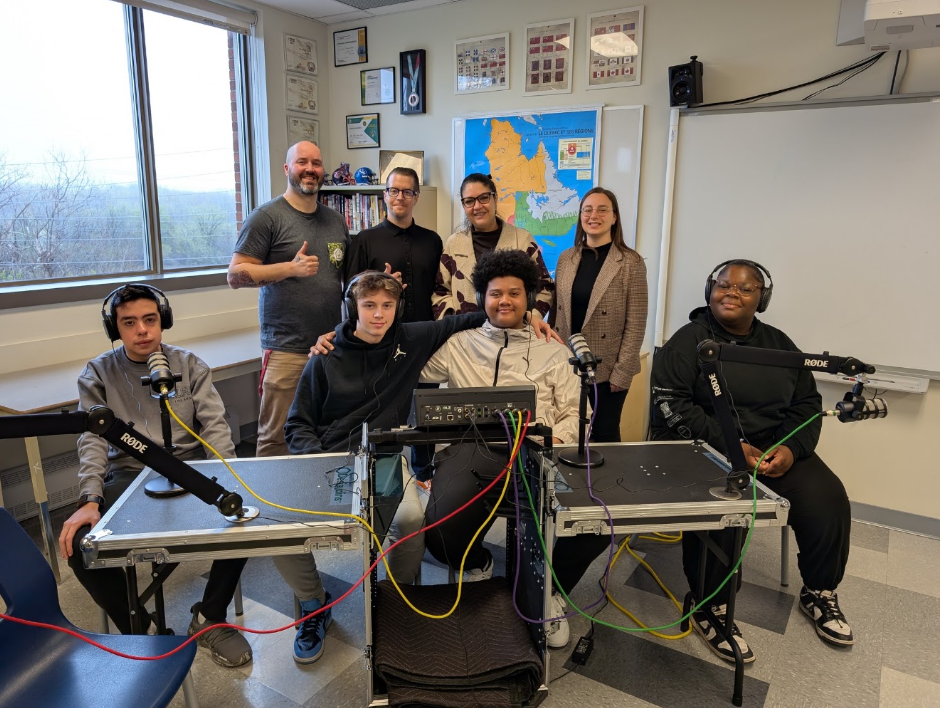 Group of eight people in a classroom recording studio with microphones and audio equipment, posing for a photo.